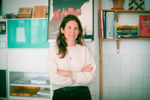 Portrait of Julia Nuño de la Rosa standing with folded arms in a modern office space with shelves, posters, and organised files in the background.