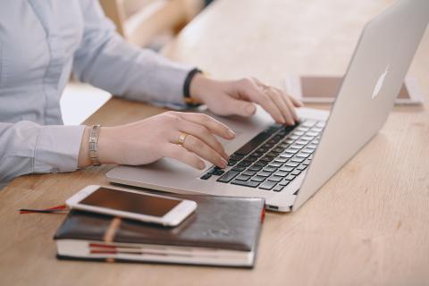 Close-up of a woman working on a laptop at a wooden desk with a smartphone and notebook beside her, symbolising online communication and digital safety.