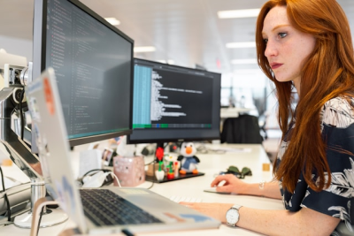 Female software developer writing and reviewing code on dual monitors in a modern office workspace.